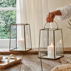 a person holding a lit candle in front of two glass lanterns on a wooden table