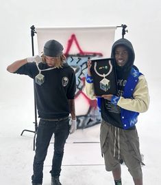 two young men standing next to each other in front of a backdrop holding up jewelry