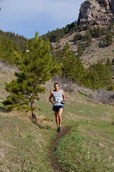 a man is running down a trail in the mountains with trees and grass on both sides