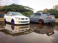 two cars parked next to each other on a wet parking lot with trees in the background