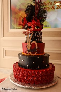 a three tiered cake decorated with red and black icing, topped with an elaborate mask