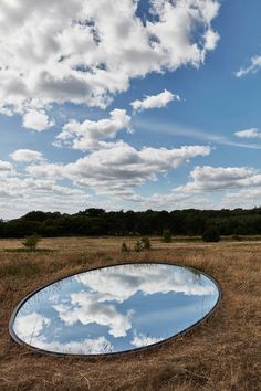 a round mirror sitting in the middle of a dry grass field under a cloudy blue sky