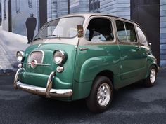 an old green car is parked in front of a wall with a painting on it