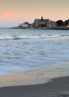 an ocean view with houses in the distance and waves crashing on the beach at sunset