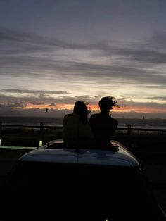 two people sitting on top of a car looking at the sunset over the ocean in front of them