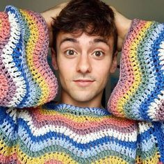 a young man with his hands on his head covering his face from the sun while wearing a multicolored crocheted sweater