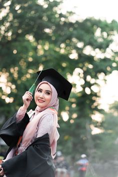 a woman wearing a graduation gown and holding a green object in her hand while standing next to a tree