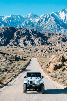 a white jeep driving down a dirt road in front of snow - capped mountain range