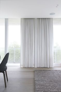 an empty living room with white curtains and grey chairs in front of a large window