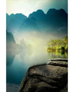 an image of mountains and water in the foggy morning time, with some trees on the shore