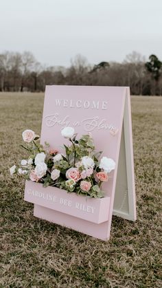 a pink welcome sign with flowers on it in the middle of a grassy field at an outdoor wedding venue