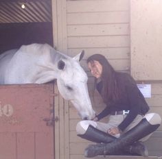 a woman sitting on the ground next to a white horse in a stable with its head sticking out