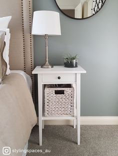 a white table with a basket under it next to a mirror and lamp on the wall