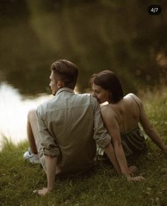 a man and woman are sitting on the grass by a river looking at each other