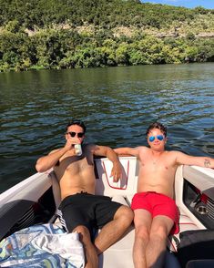 two men sitting in the back of a boat on a body of water with trees in the background