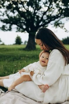 a woman holding a baby in her lap while sitting on the ground next to a tree