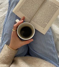 a person laying in bed reading a book and holding a cup of coffee with both hands