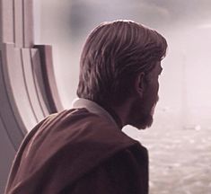 a man with long hair and beard standing next to a window looking out at the water