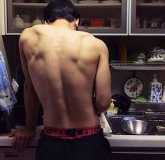 a man with no shirt standing in front of a kitchen sink and looking at dishes on the counter