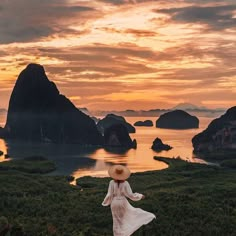 a woman in a white dress and hat standing on top of a hill looking at the ocean