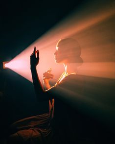 a woman is sitting in the dark with her hand up and light coming from behind her