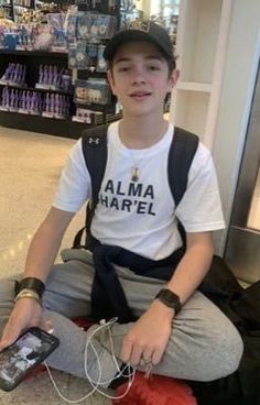 a young man sitting on the ground holding a cell phone and wearing a white t - shirt