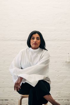 a woman sitting on top of a chair in front of a white brick wall and wearing black pants