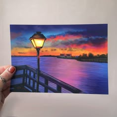 a person holding up a painting of a light post by the water at sunset or sunrise