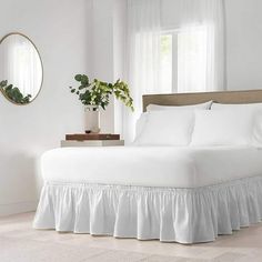 a white bed in a bedroom next to a window with curtains and a mirror on the wall