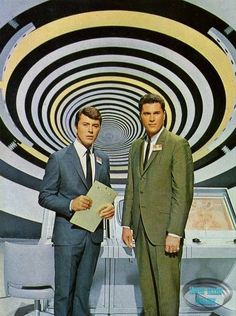 two men standing next to each other in front of a spiral background with an open book