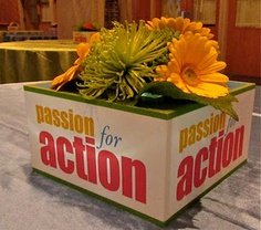 an action sign with flowers in it sitting on a table at a convention or event