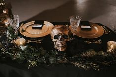 a table topped with gold and black plates covered in greenery next to a skull head