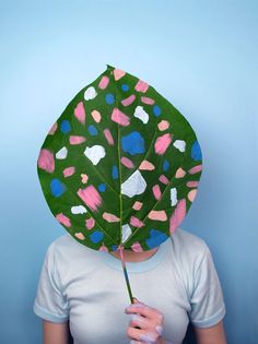 a woman holding a large leaf over her face with paint splattered on it