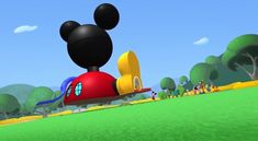 mickey mouse flying in the air over a green field with trees and people on it