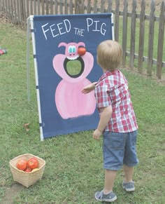 a young boy standing in front of a feed the pig sign with apples on it