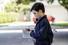 a young boy writing on a piece of paper while standing in front of a tree