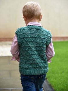 a little boy wearing a green knitted sweater vest and jeans walking down a sidewalk