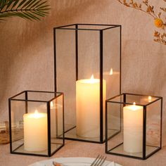 three square glass candle holders with lit candles in them on a table next to a plate