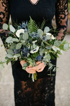 a woman in a black dress holding a bouquet of greenery and blue thistles