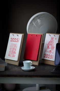 three calendars sitting on top of a wooden table next to a laptop and coffee cup