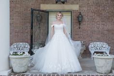 a woman standing in front of a door wearing a white wedding dress with an off the shoulder neckline