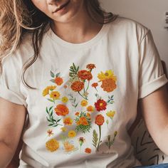 "Marigold Botanical Shirt 'Like a Well Watered Garden' This marigold botanical shirt is perfect for anyone who loves nature and wants to express their love of plants. This botanical shirt is soft and cozy featuring a pretty wildflower design. The shirt is machine-washable and fits comfortably, whether you're wearing it for a day at the park or lounging around the house. Give the gift of comfort and style with this marigold botanical shirt. The inspiration is from the scripture text Isaiah 58:11 Fall Yellow Shirt With Floral Print, Orange Floral Print Shirt For Spring, Orange Floral Embroidery Short Sleeve Tops, Orange Short Sleeve Tops With Floral Embroidery, Orange Floral Embroidered Short Sleeve Tops, Orange Cotton Floral Print Shirt, Orange Floral Print Cotton Shirt, Yellow Cotton Shirt With Floral Print, Yellow Cotton Floral Print Shirt