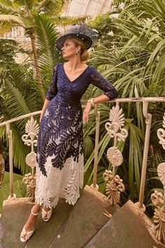 a woman in a blue dress and hat standing on a balcony with palm trees behind her