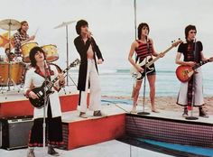 the rolling stones on stage with their guitars in front of an ocean and sky background