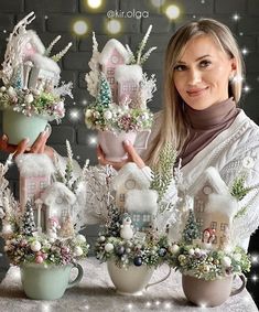 a woman is holding three cups with small houses in them and christmas decorations on the table