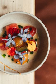 a white bowl filled with fruit and flowers on top of a wooden table next to a spoon