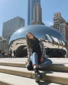 a woman sitting on the steps in front of a large metal object with buildings behind her