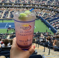 a person holding up a drink in front of a tennis stadium filled with people watching