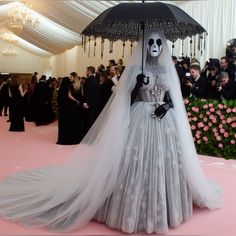 a woman in a wedding dress and veil with an umbrella on the pink carpet, surrounded by people