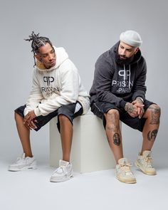 two young men sitting next to each other on top of a white pedestal wearing hoodies